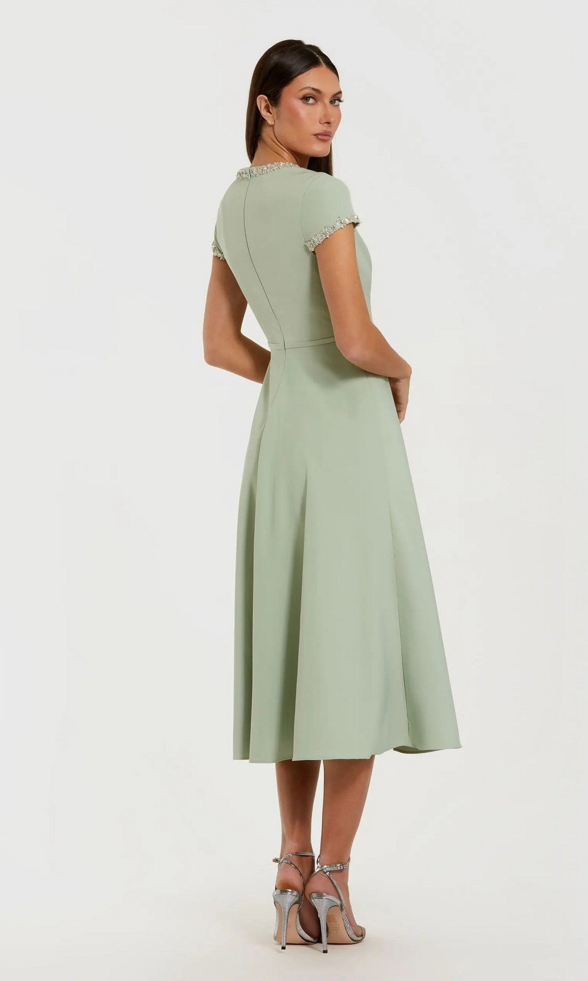 A woman stands with her back to the camera, wearing the Midi Party Dress: Mac Duggal 56242—a light green, short-sleeved, knee-length dress featuring beaded trim—paired with silver high-heeled sandals against a plain background.